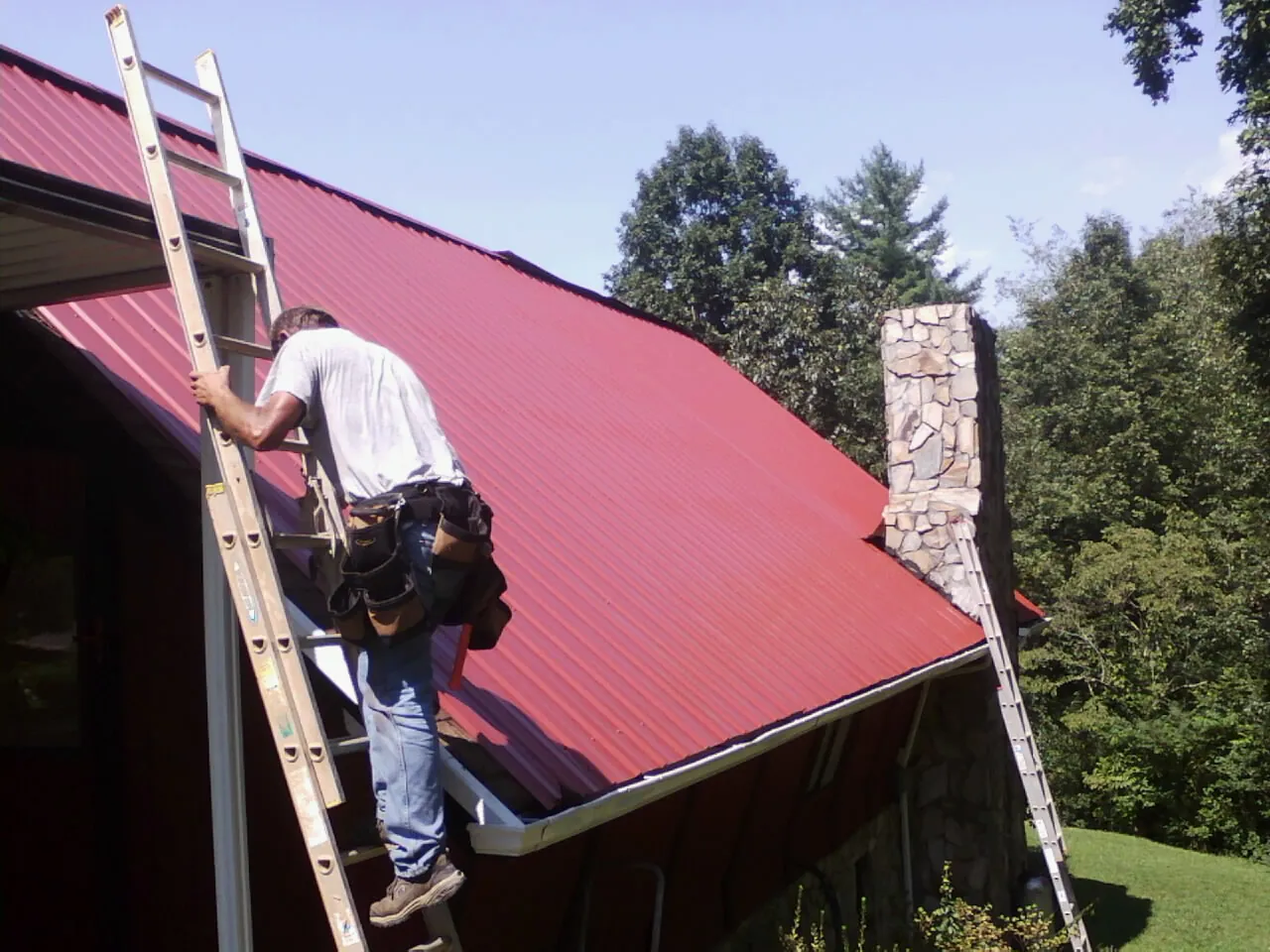 Roofer on a ladder beside a red metal roof and stone chimney — Soffit & Fascia Repair by Roofing Brothers in Silverdale